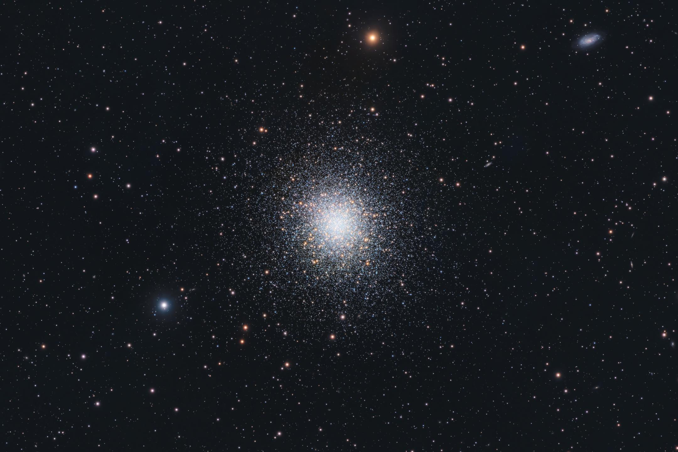 A dense ball of tens of thousands of stars, blue-white and unresolved at the core, thinning into separately visible points towards its edges, set in a sparse field of orange and blue foreground stars; a small edge-on galaxy sits near the top right corner.