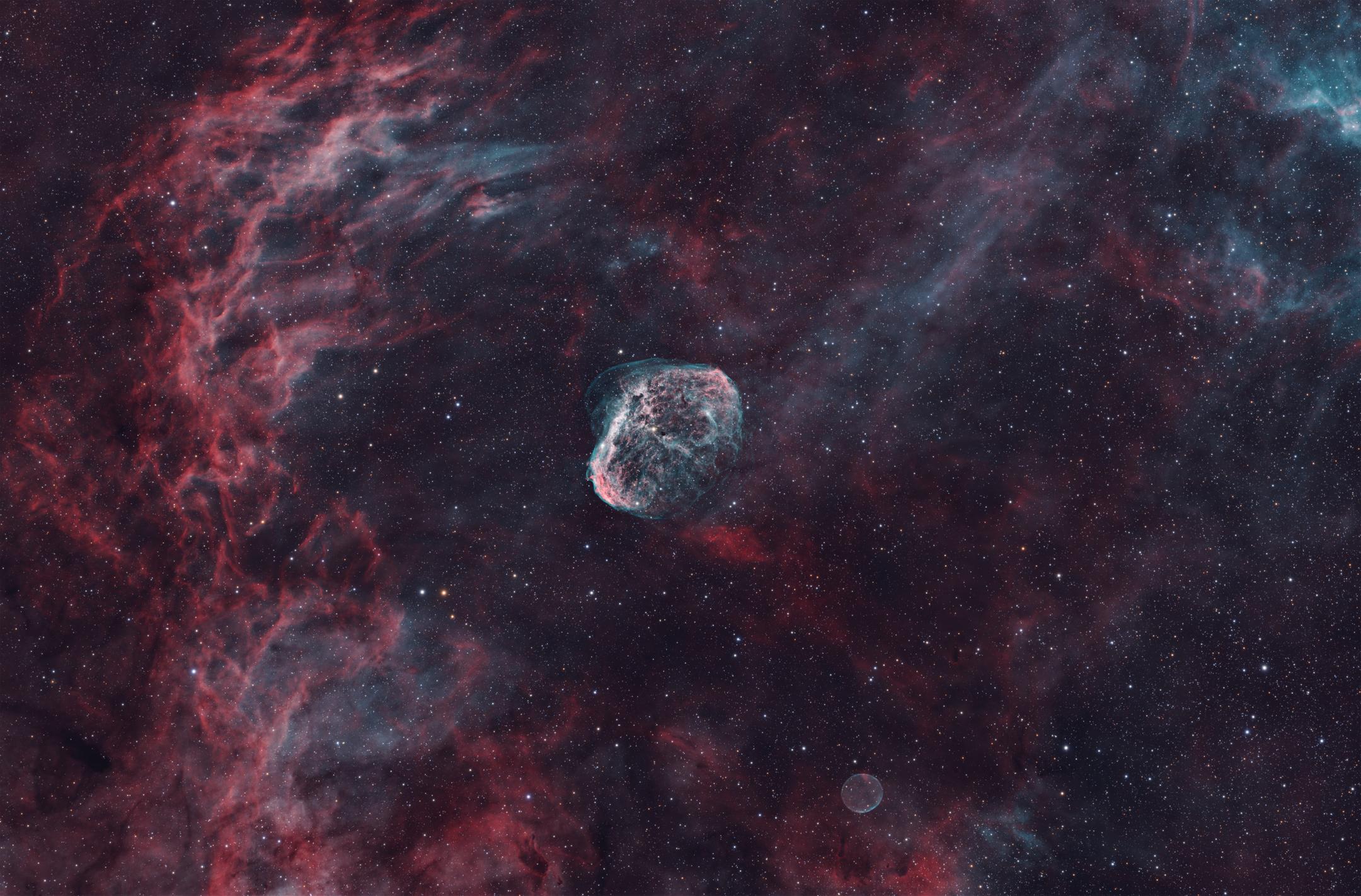 A compact, crumpled shell of gas near the centre, teal within and rimmed in red, set against sweeping filaments of red and blue-green; a small, much fainter ring floats near the bottom of the frame.