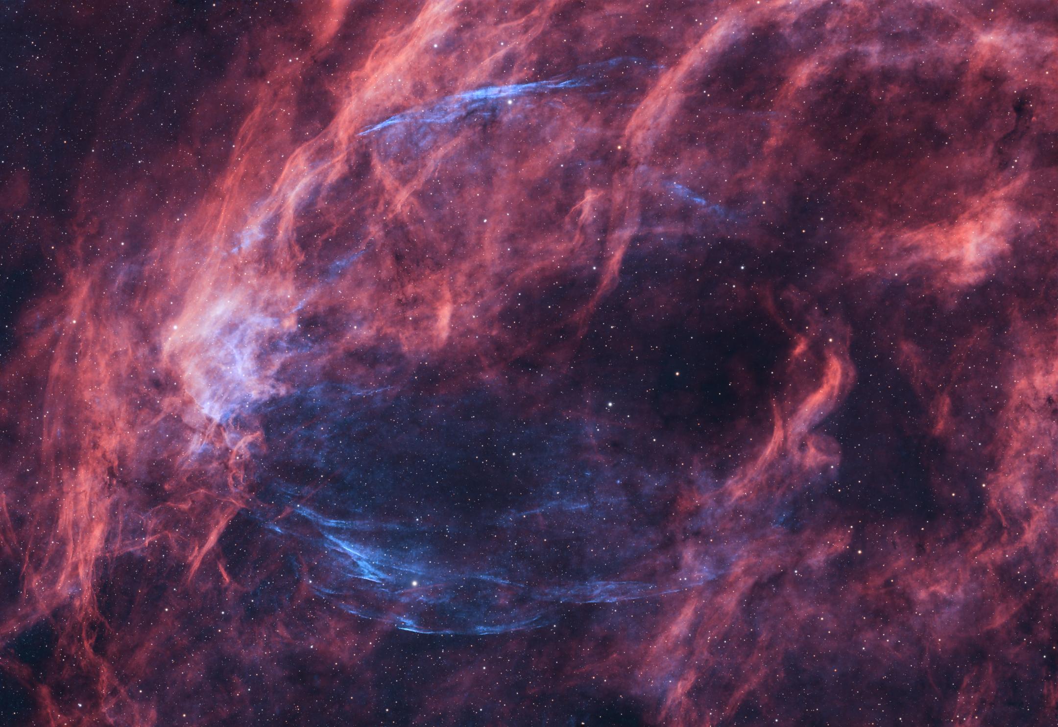 Thick red-pink filaments filling the frame, with a much fainter blue arc looping through the middle of them and tracing out the edge of a large, roughly circular shell.